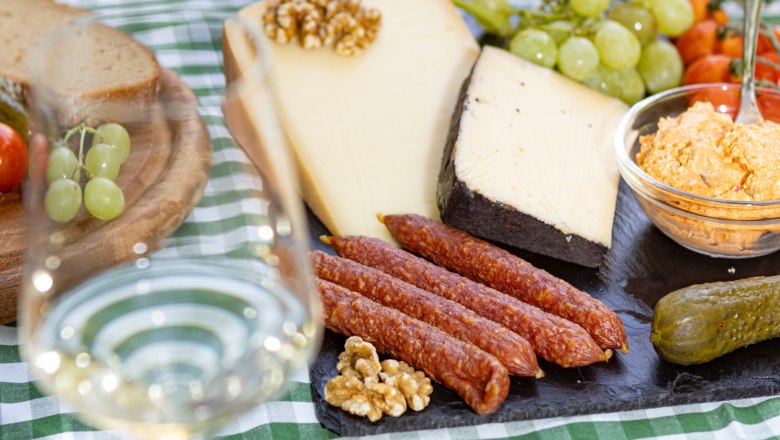 Heurigenjause, © Weinviertel Tourismus GmbH / Schwarz-König Picknicktisch mit Käse, Wurst, Brot, Weintrauben und einem Glas Weißwein.