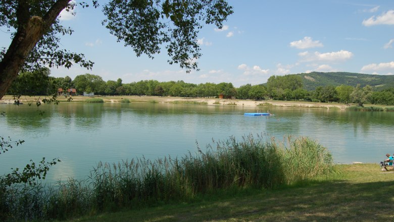 Seeschlacht, © Gemeinde Langenzersdorf Ein ruhiger See mit Bäumen und einem Steg im Hintergrund, umgeben von grüner Landschaft.