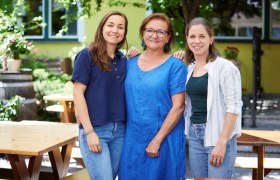 Familie Schmidt, © Ben Leitner Drei Frauen stehen lächelnd in einem Garten mit Holztischen und Pflanzen.