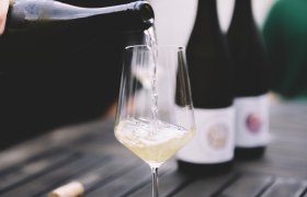 Wein einschenken, © Niederösterreich Werbung / Mara Hohla Ein Glas Weißwein wird auf einem Tisch eingeschenkt, mit zwei Weinflaschen im Hintergrund.