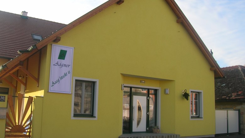 Außenaufnahme, © Fam Aigner Gelbes Gebäude mit einem Schild "Aigner Ausg'steckt is" an der Fassade.