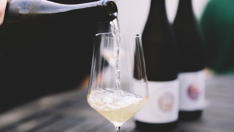 Wein einschenken, © Niederösterreich Werbung / Mara Hohla Ein Glas Weißwein wird auf einem Tisch eingeschenkt, mit zwei Weinflaschen im Hintergrund.