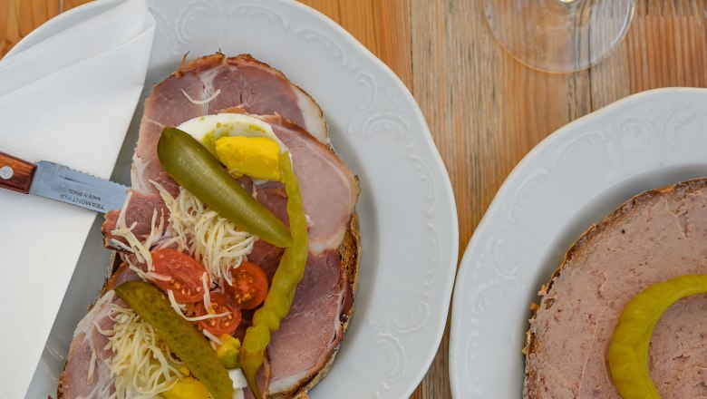 Heurigenjause, © Weinviertel Tourismus GmbH / Lisa Sedlatschek Zwei Teller mit belegten Broten und zwei Gläser Weißwein auf einem Holztisch.