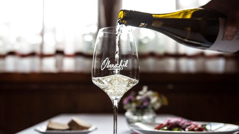 Heuriger Oberschil, © Astrid Bartl Ein Glas Weißwein wird eingeschenkt, im Hintergrund ein gedeckter Tisch mit Blumen.