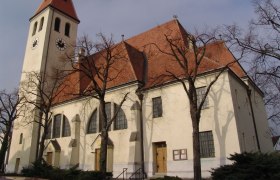 Kirche in Enzersfeld, © Pfarre Enzersfeld Kirche mit Turm und roten Ziegeldach, umgeben von kahlen Bäumen.
