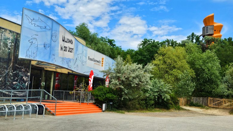 Herzlich Willkommen, © Fossilienwelt Eingang der Fossilienwelt mit einem großen Banner und Bäumen im Hintergrund.