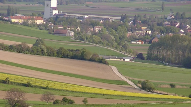 Landschaft mit Feldern und einem Dorf im Hintergrund, dominiert von einem gro&szlig;en wei&szlig;en Geb&auml;ude mit dunklem Dach.