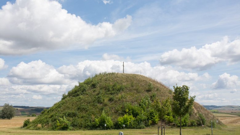 Ein grasbewachsener H&uuml;gel mit einem Kreuz auf der Spitze unter einem bew&ouml;lkten Himmel.
