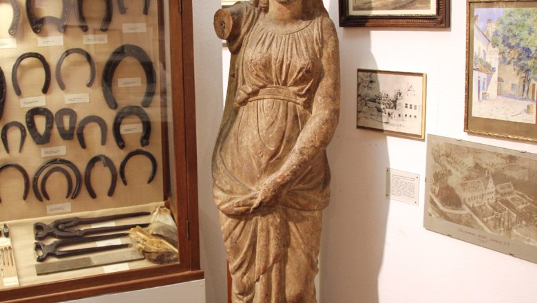 Innenansicht eines Museums mit antiker Statue, Hufeisen in einer Vitrine und historischen Bildern an der Wand.