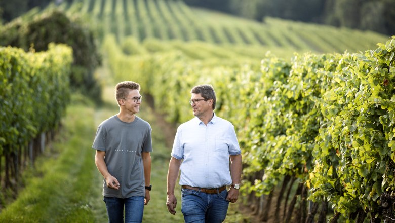 Zwei Männer gehen lächelnd durch einen Weinberg.