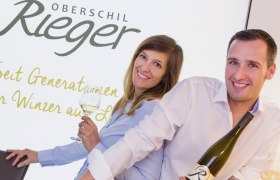 Zwei Personen lächeln und halten Weinflaschen vor einem Schild mit der Aufschrift 'Oberschil Rieger'.
