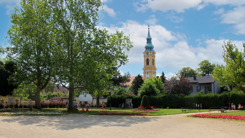 Ein Park mit Bäumen, Blumenbeeten und einem Kirchturm im Hintergrund.