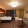 Modern eingerichtetes Hotelzimmer mit Doppelbett, Sofa, zwei Sesseln und einem kleinen Tisch. Holzboden und warme Beleuchtung.